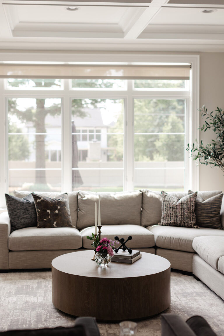 A modern living room with a large beige sectional sofa, patterned pillows, a round wooden coffee table with decor, and large windows overlooking a green yard.
