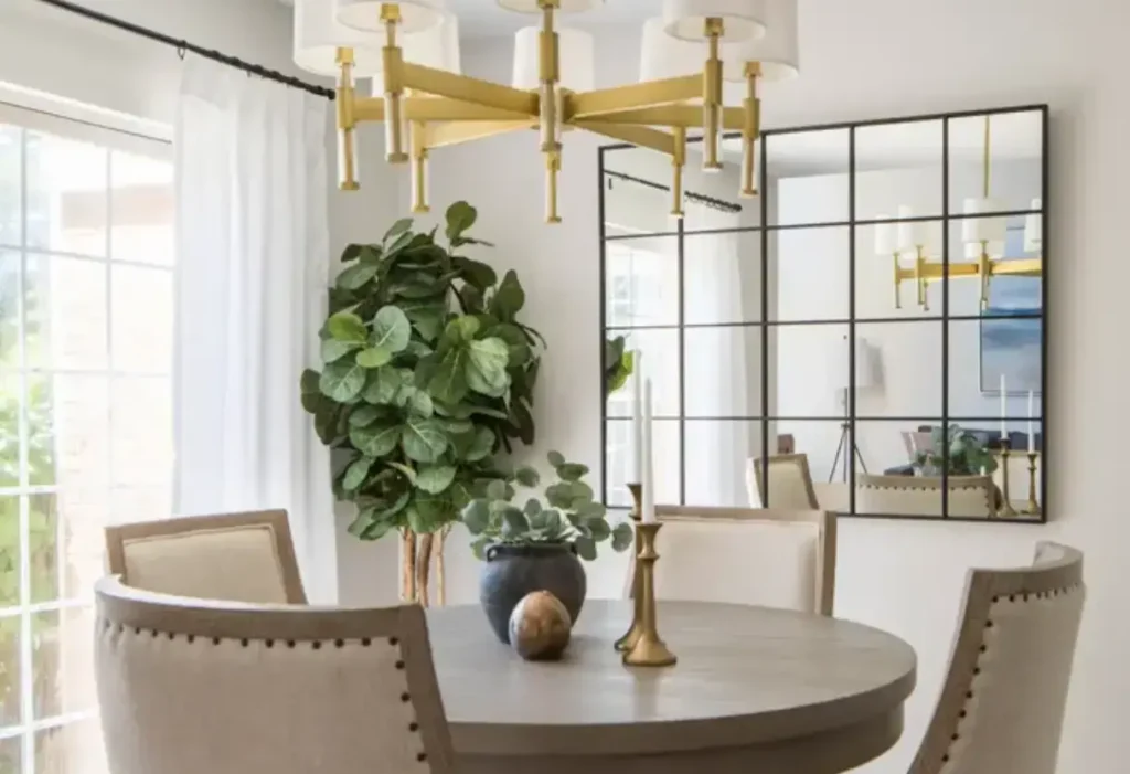 A round dining table with beige chairs, a potted plant, a gold chandelier, and a large grid mirror on the wall in a bright, modern dining room.