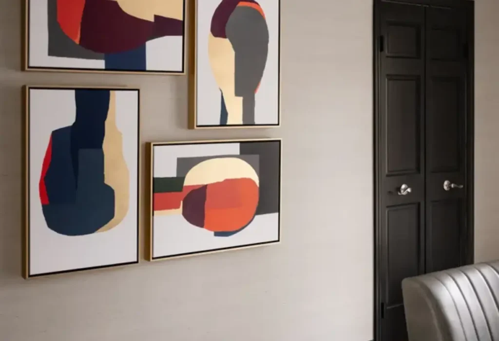 Three abstract paintings with bold geometric shapes and muted colors hang on a light wall near a black door and a gray chair.