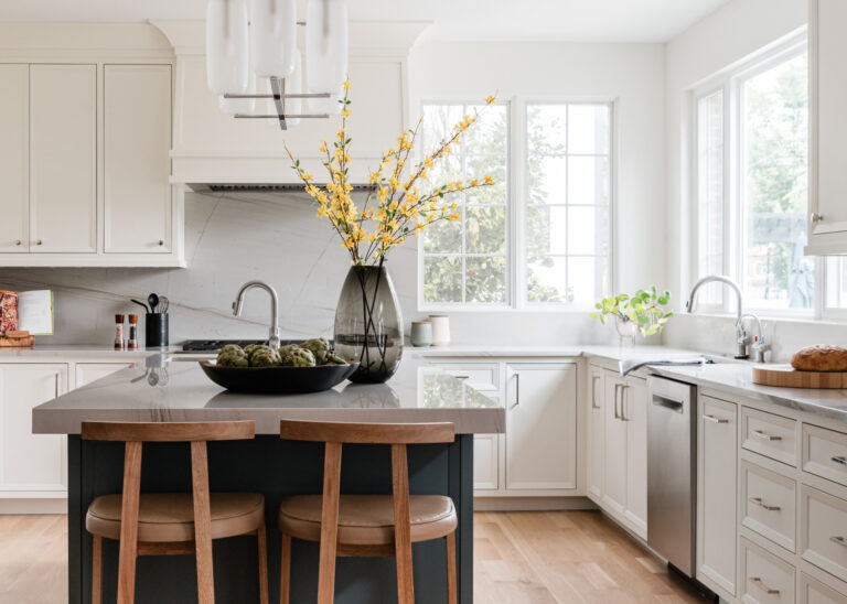Modern kitchen with white cabinets, a large island with two wooden stools, a vase of yellow flowers, and natural light coming through two large windows.