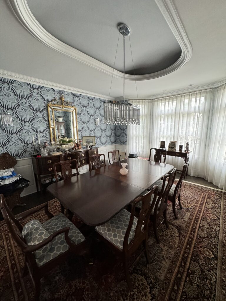 A formal dining room with a large wooden table, ten chairs, a patterned rug, chandelier, sideboard with a mirror, and patterned wallpaper, lit by natural light from large windows.