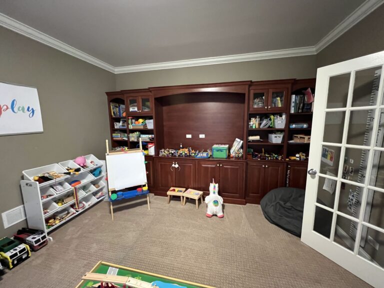 A playroom with brown built-in shelves and cabinets filled with toys, a toy storage shelf, an easel, a small table, and a plush unicorn on a carpeted floor.