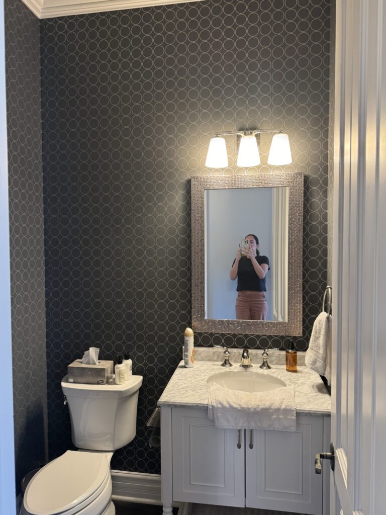 Person taking a mirror selfie in a modern bathroom with dark patterned wallpaper, a white toilet, and a white sink vanity under a lit three-bulb fixture.