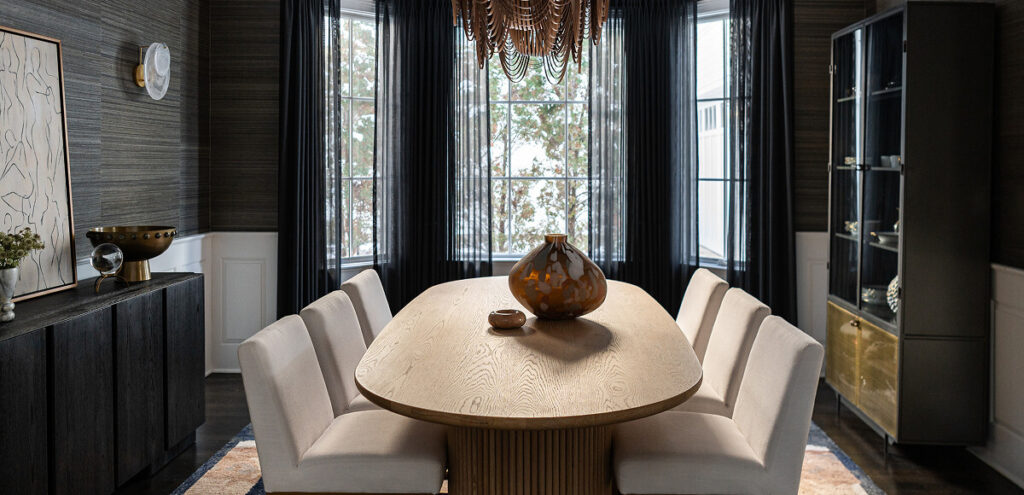 Modern dining room with a large wooden table, eight white chairs, a decorative vase centerpiece, dark cabinets, and a window with sheer black curtains.