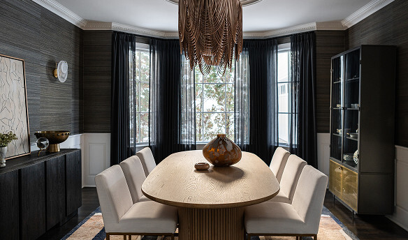 Modern dining room with a light wood oval table, eight upholstered chairs, a glass cabinet, abstract wall art, and a large, intricate chandelier below a bay window with dark curtains.