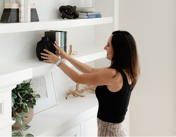 Woman arranging a black vase on a white bookshelf with books, decor items, and a framed photo on the lower shelf.