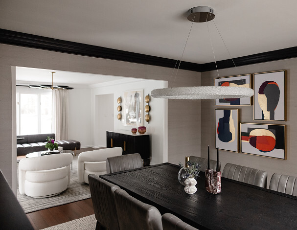 Modern dining room with a dark wood table and gray chairs, abstract art on the wall, and a circular light fixture; living room with white chairs and wall decor visible in the background.