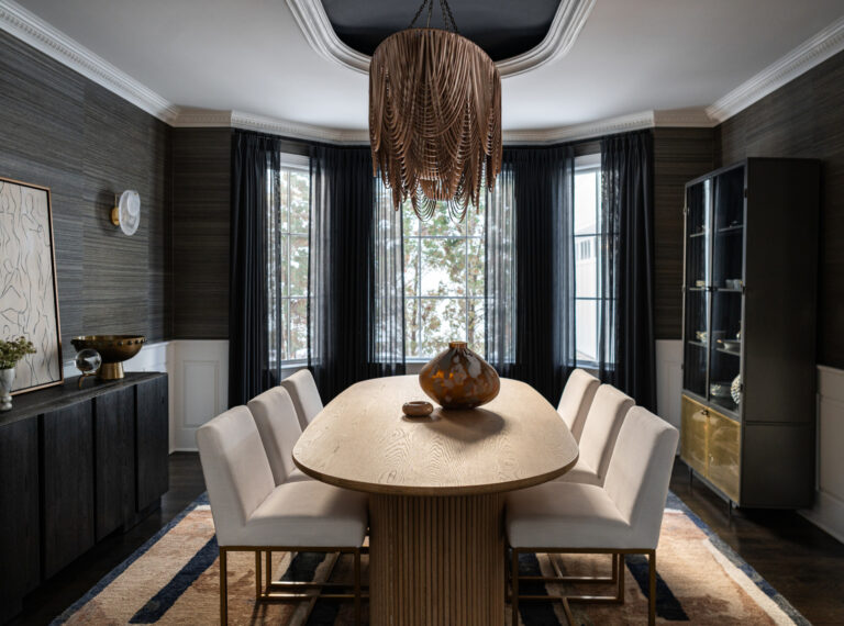 Modern dining room with a wooden oval table, eight white cushioned chairs, a large decorative vase, dark curtains, and a unique chandelier hanging from the ceiling.