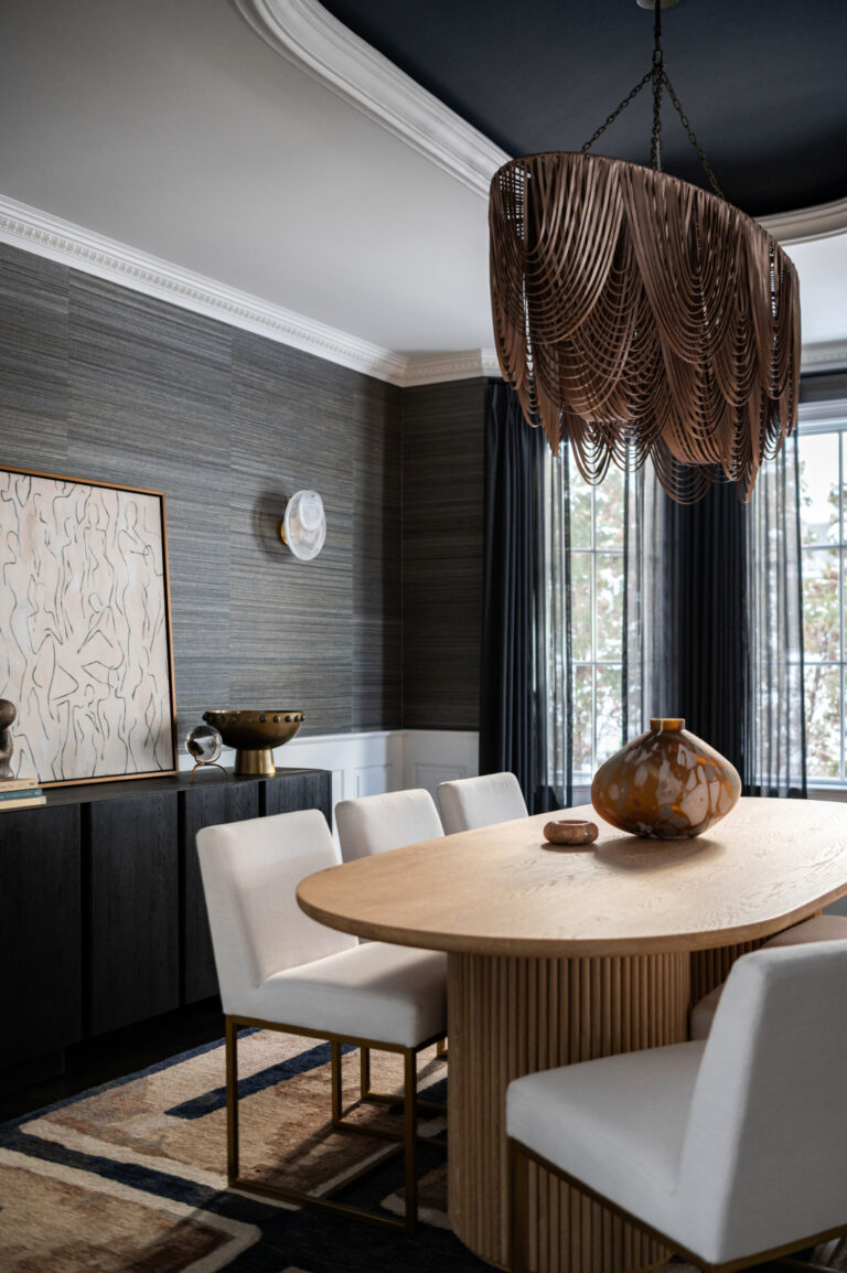 A modern dining room with a wooden oval table, six white chairs, a large decorative chandelier, and contemporary art on the wall. Natural light enters through tall windows with dark curtains.