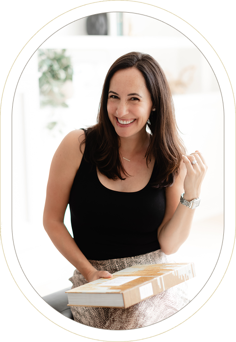 Smiling woman with long brown hair in a black sleeveless top sits holding a closed cardboard box on her lap in a bright indoor setting.