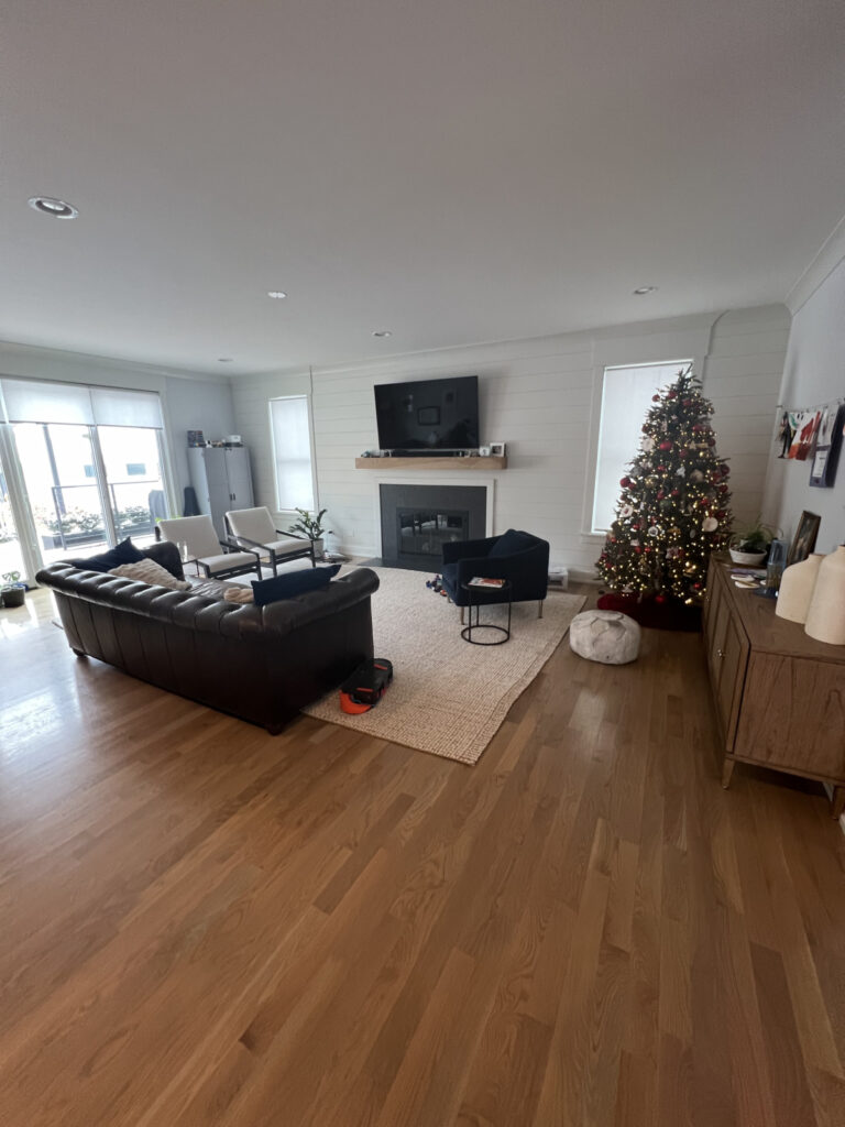 A living room with wooden floors, a brown leather sofa, a TV above a fireplace, a decorated Christmas tree, and natural light coming through large windows.