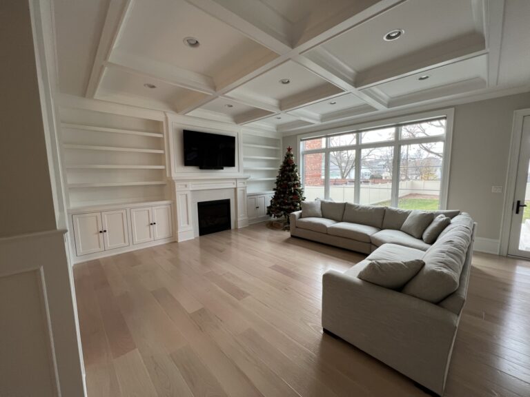 A spacious living room with light wood floors, built-in shelves, a fireplace with a TV above, a Christmas tree, and a large sectional sofa near a window.