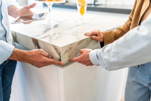 Three people stand around a marble kitchen countertop, examining the edge; one person holds a tablet while the others point at the counter’s corner.