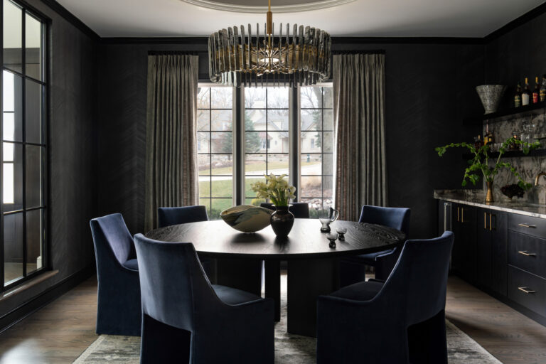 A modern dining room with dark walls, a round black table, six blue chairs, a chandelier, large windows with curtains, and a sideboard with plants and glassware.