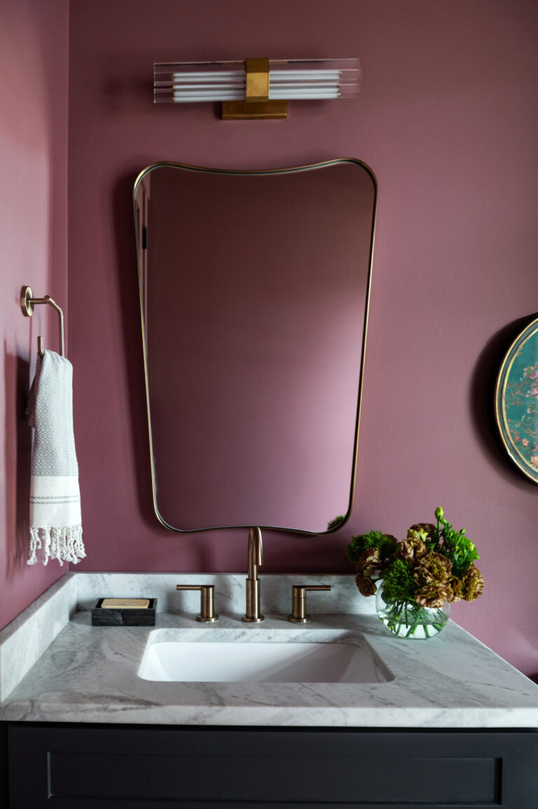 A bathroom vanity with a marble countertop, gold fixtures, an oval mirror, a towel on a ring, and a vase of flowers against mauve walls.