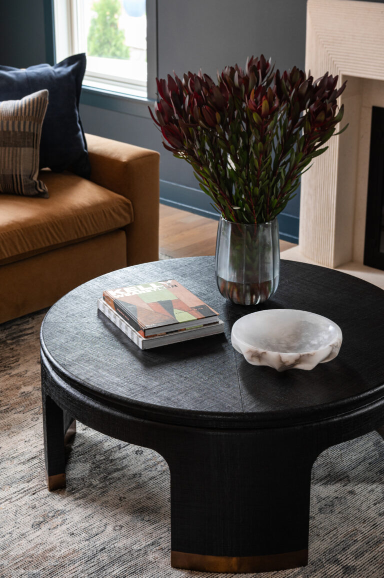 A round black coffee table with a glass vase of dark red flowers, a modern book, and a white decorative bowl, in a living room with a tan sofa and gray walls.