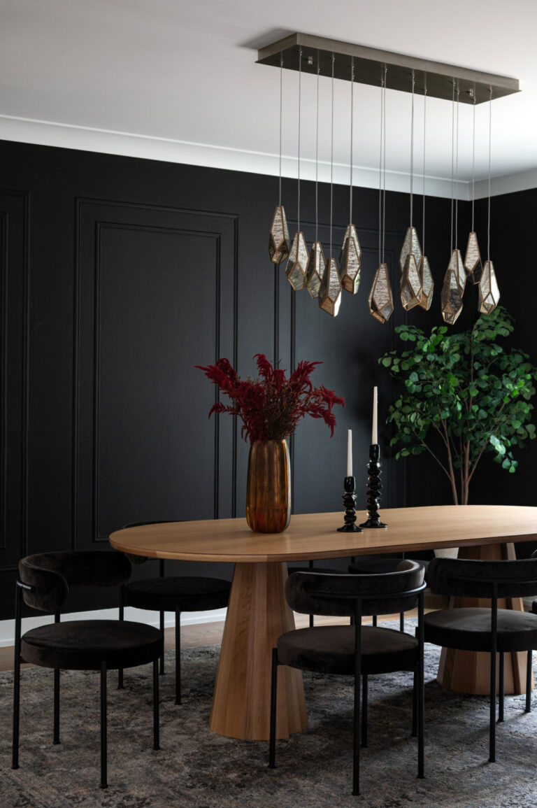 A modern dining room with a wooden oval table, black chairs, a gold vase with red flowers, two black candlesticks, a geometric chandelier, black walls, and a green plant in the corner.