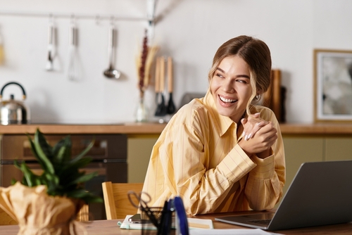 Woman sitting at a kitchen table, smiling and looking to the side, with a laptop open in front of her and kitchen utensils visible in the background.