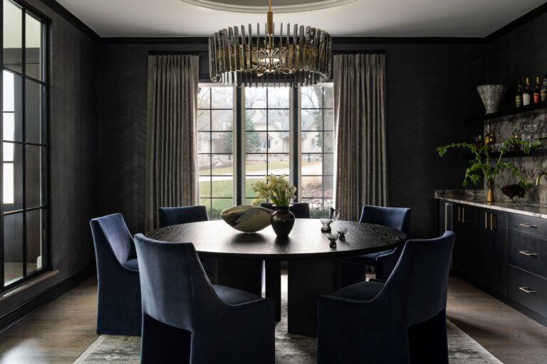 A modern dining room with a round table, six blue velvet chairs, large windows with beige curtains, dark walls, and a contemporary chandelier overhead.