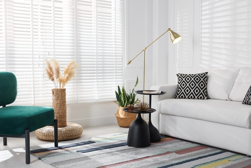 Modern living room with a white sofa, geometric cushions, green chair, houseplants, and a gold floor lamp on a colorful striped rug near large windows with blinds.