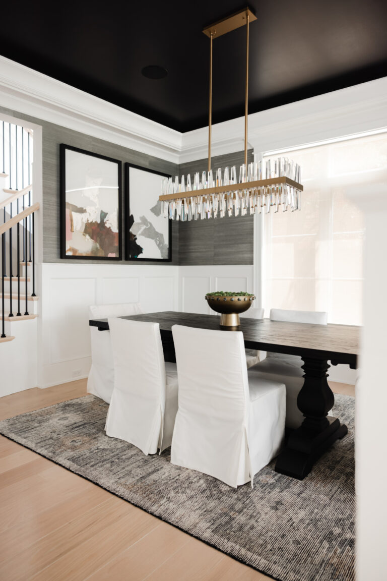 Modern dining room with a dark wood table, six white upholstered chairs, abstract wall art, a large chandelier, and a gray rug on light wood flooring.