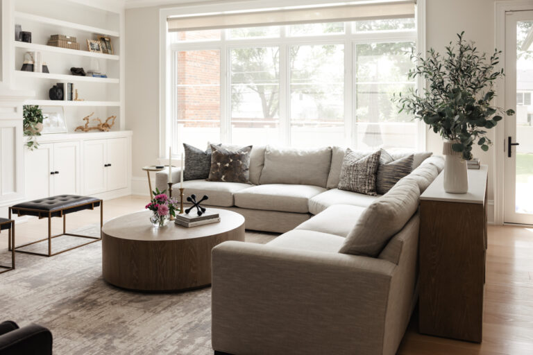 Modern living room with a large L-shaped sofa, round coffee table, decorative pillows, built-in shelves, and large windows letting in natural light.