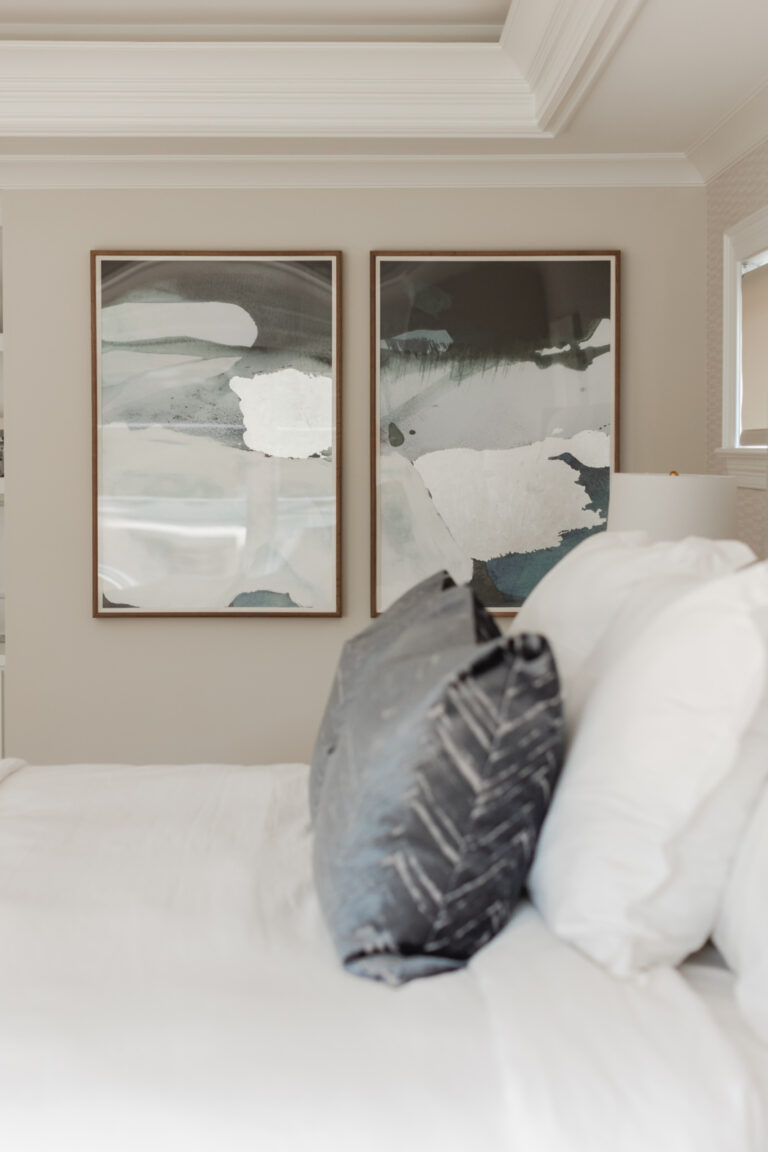 A neatly made bed with white linens and a dark patterned pillow, positioned beside a wall featuring two large abstract paintings in neutral tones.