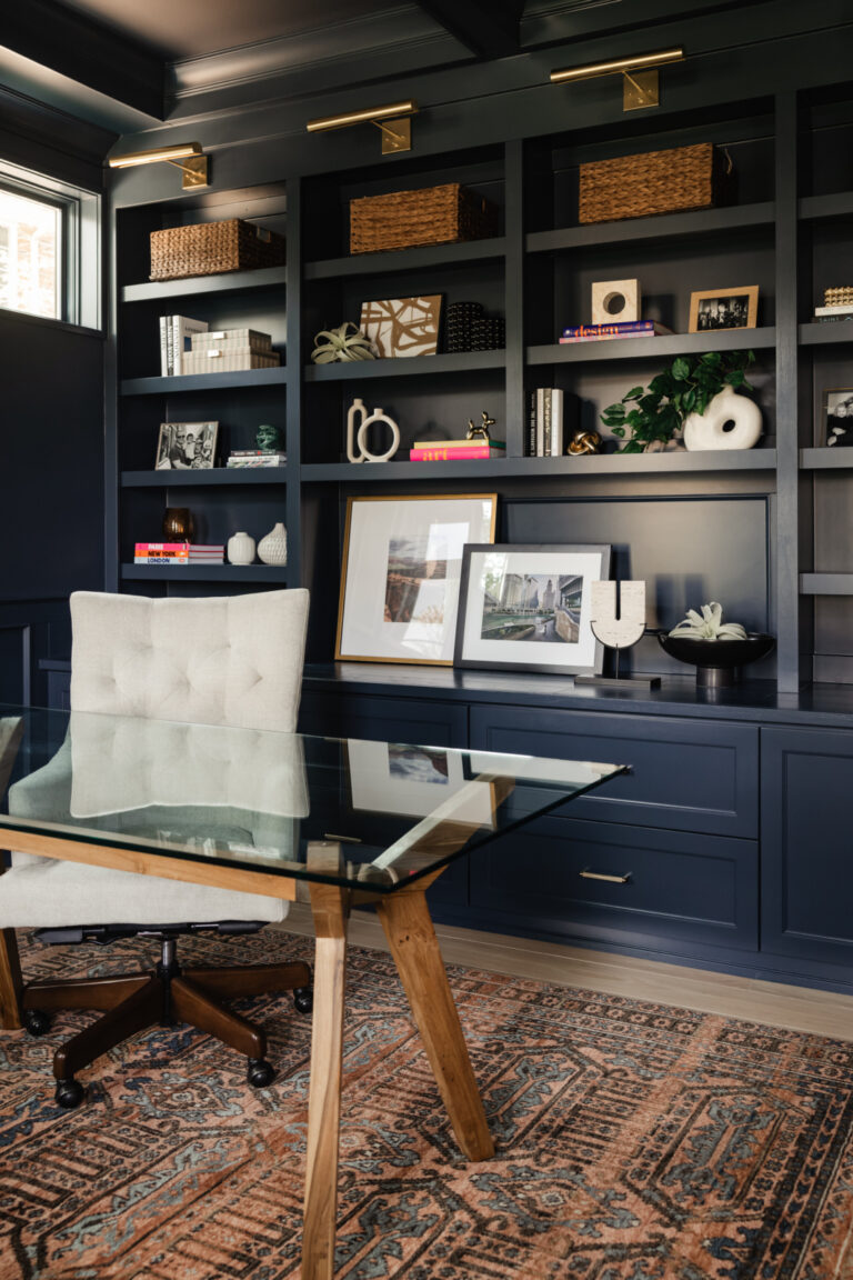 A modern home office with a glass desk, upholstered chair, and built-in navy shelves displaying books, baskets, framed photos, and decor items on a patterned rug.