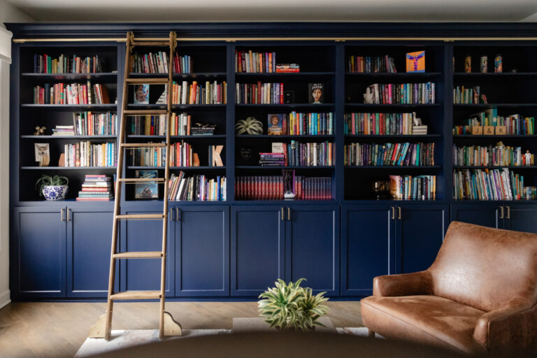 A large built-in bookshelf with blue cabinets, a rolling ladder, and filled with books and decor items, next to a brown leather chair and a small plant on the floor.